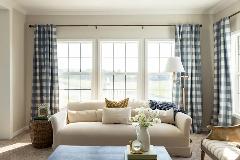 Three large windows are framed by blue checkered curtains and are behind a cream couch in a living room.