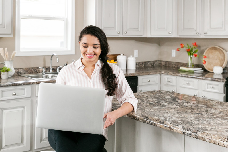 Woman with dark hair sits in a kitchen  at a white kitchen island with marble countertops while looking at the screen of a silver laptop.