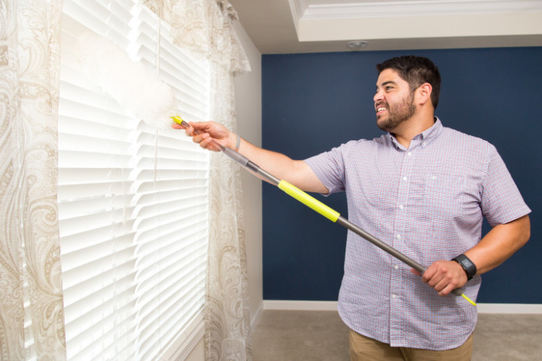 A young man using a duster to dust window blinds in a bedroom with a blue accent wall.