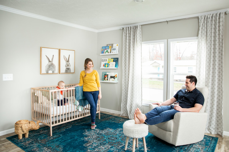 Woman standing next to crib with child and man sitting in chair in manufactured home nursery.