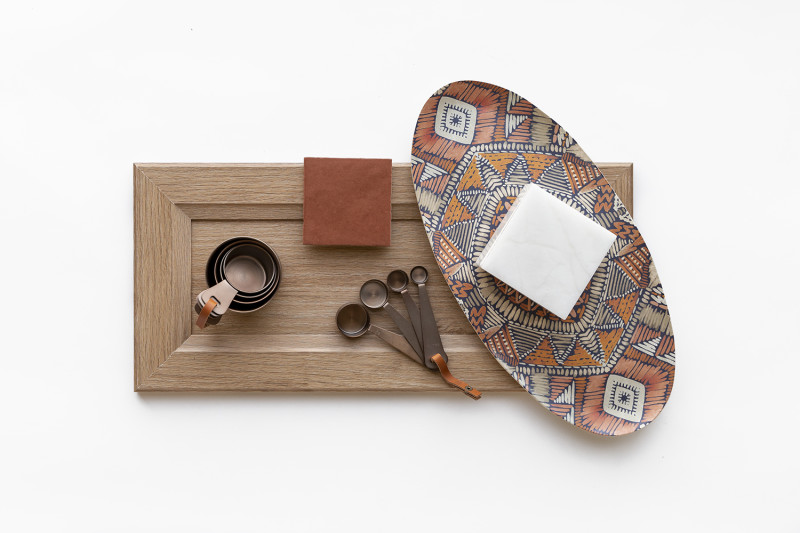 A flat lay of earthy tones and a light wood cabinet, patterned dish, measuring spoons and cups and a terracotta tile