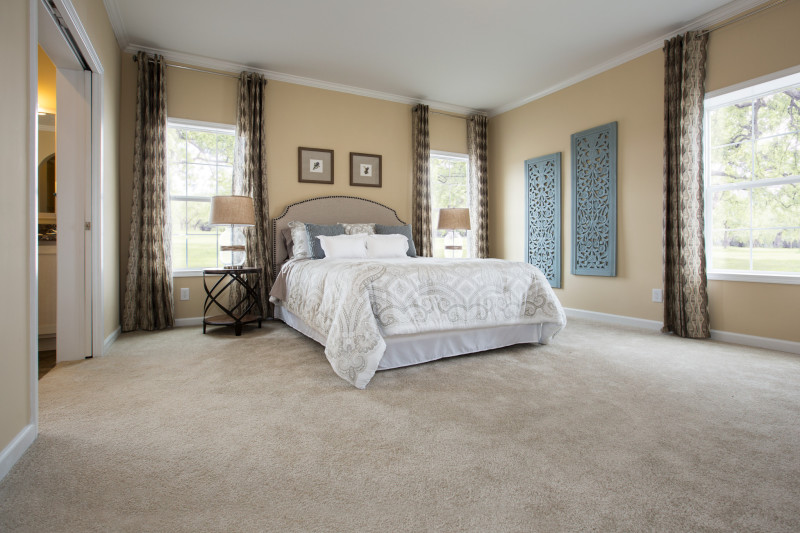 View is of a tan bedroom with large windows with dark drapes hanging on them and tons of natural light streaming in.