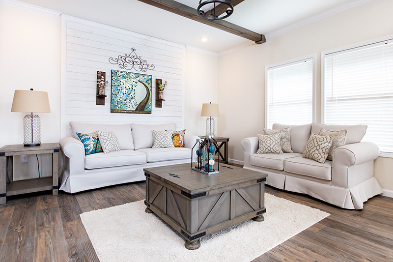 Living room of the Southern Charm manufactured home with shiplap accent wall, ceiling beams, windows, 2 couches and wooden coffee table and end table.