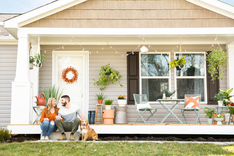 Exterior of a CrossMod home with wood shingles, tan siding and white porch and trim, with plants and tables and chairs, and a couple with a dog sitting on the front step.