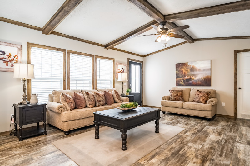 Living room of a single wide manufactured home with medium wood trim, ceiling beams and fan, beige couches and dark wood tables, and weathered wood-style flooring.