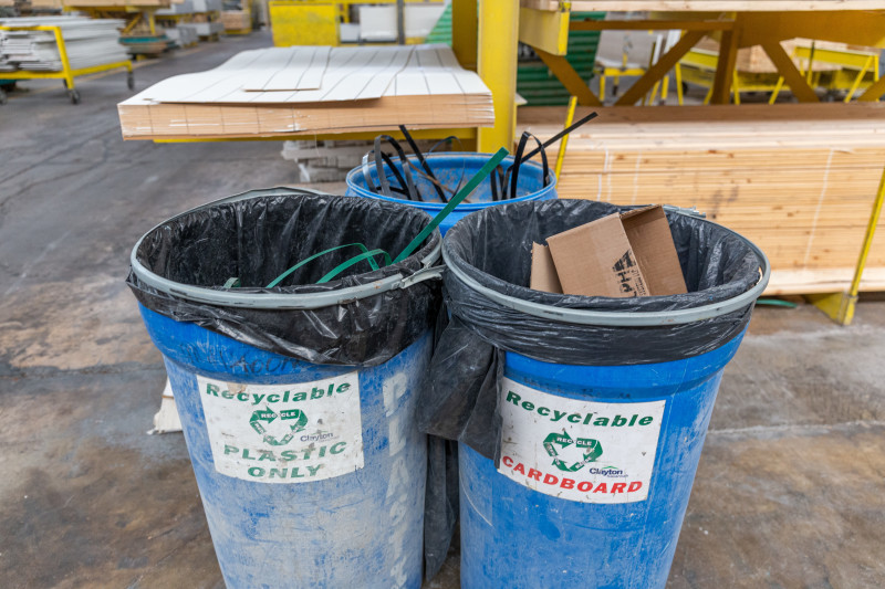 Three blue trash cans for recycling plastic and cardboard inside of a manufactured home building facility, with shelves of wood in the background and a concrete floor.