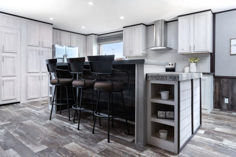 Angled view of a kitchen with a large island. There are brown and black chairs pulled up to it for dining. Kitchen has tons of white cabinetry. There are stainless steel appliances as well as a modern kitchen hood that has a clear glass cover.