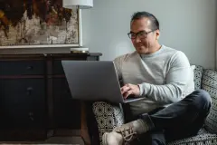 Man with glasses sitting on a chair with his legs crossed working on his laptop.