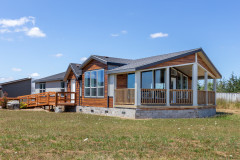 A manufactured home with warm wood and white siding, large windows and a prow porch under a clear blue sky.