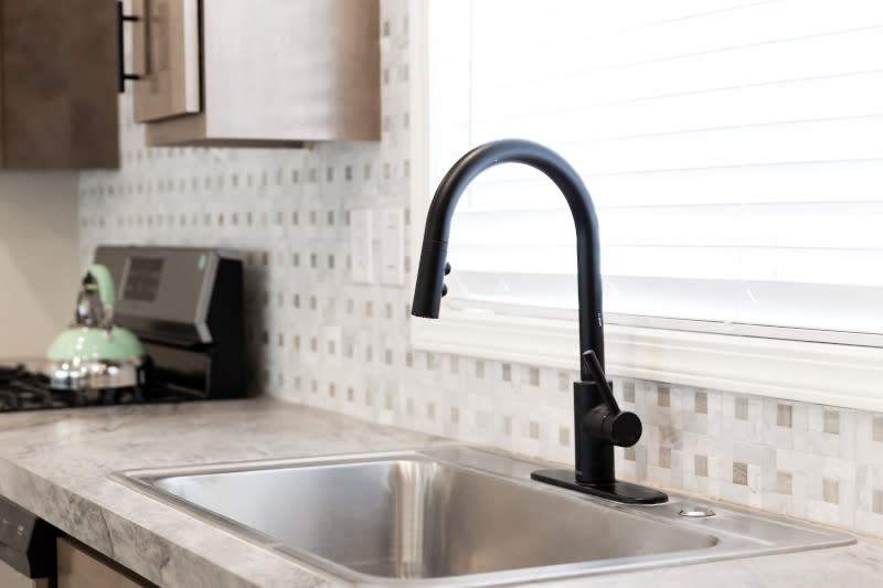 Black Pfister faucet in the kitchen island of a manufactured home kitchen featuring tile backsplash, black and silver stove and a window behind the sink.