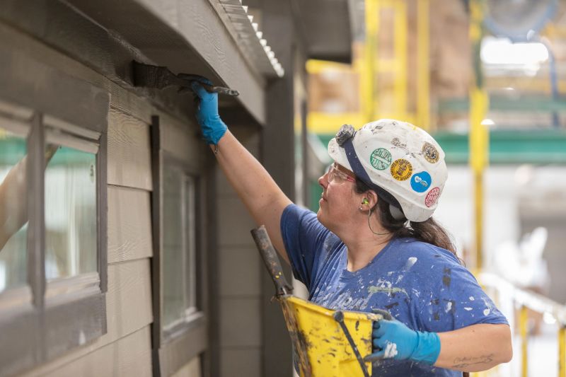 Woman wearing a hard hat and safety classes paints the exterior of a Clayton manufactured home inside a building facility.