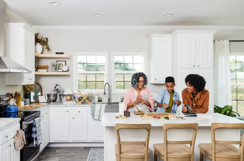 In a white and marble kitchen, a grandmother, daughter and grandson prepare cookies to back at the island.