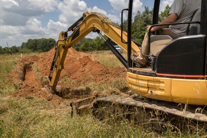 A man operates a small bulldozer to move a pile of dirt in the grass at a manufactured home site, with trees and a sky in the background.