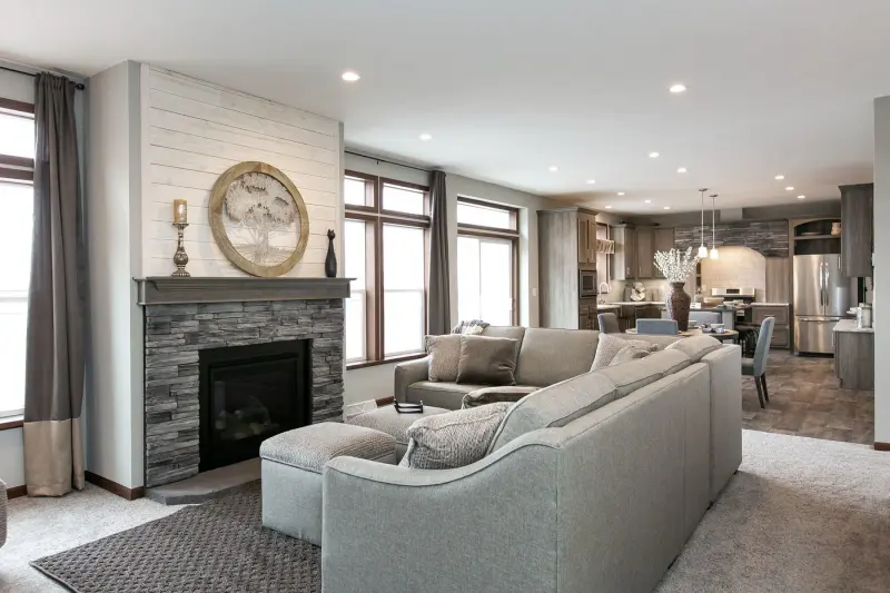 A living room with a gray couch in front of a stone fireplace is at the forefront, with a dining room and kitchen with an arched stone hood range in the background.