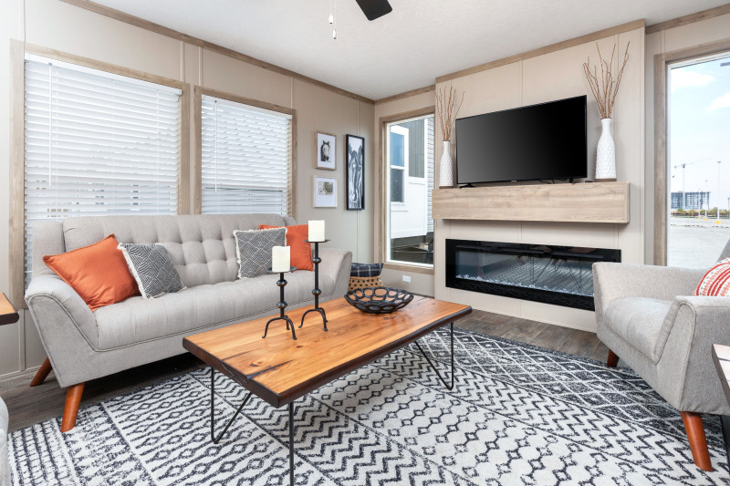 The den in this manufactured home has tons of natural light with four large windows. There is a modern fireplace with a flatscreen on the mantle. Furniture is gray with orange accents.