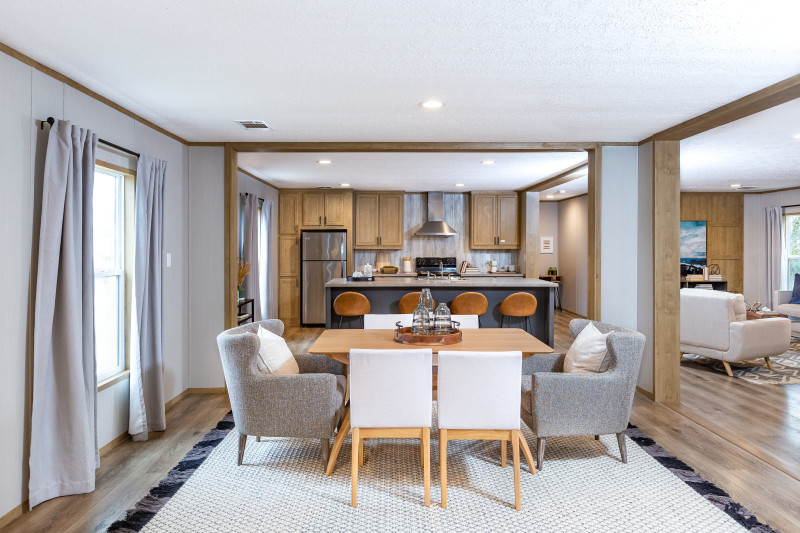 A manufactured home’s dining room with a wood table surrounded by white chairs and two gray chairs on the ends with white pillows; the dining area has an entryway to the kitchen with a blue island and another entryway to the living room with a cream chair.