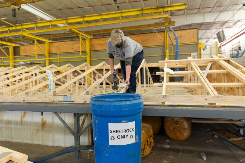 A Clayton facility worker stands on the wooden frame of a roof while tossing a circle of sheetrock in a recycling bin. 