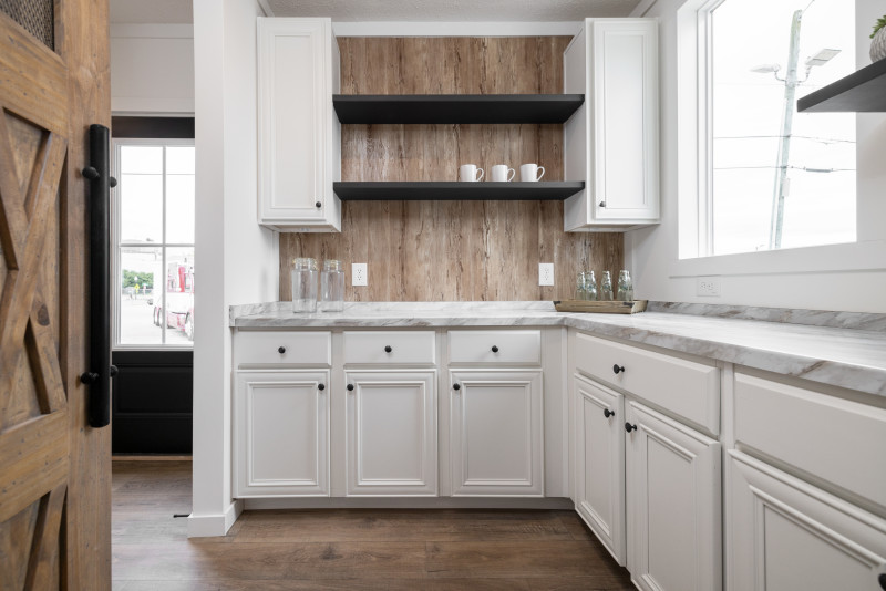 An open-door entry to the walk-in pantry in home. The cabinets are white, with wooden backsplash and black open shelving.