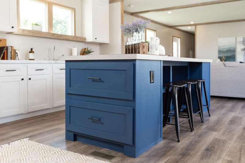 Kitchen of a manufactured home with a blue island with drawers and seating, weathered wood floor, white cabinets and light wood trim.
