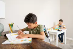 An older child writes letters at a desk in a school notebook while a younger child plays with blocks on a white table in the background.