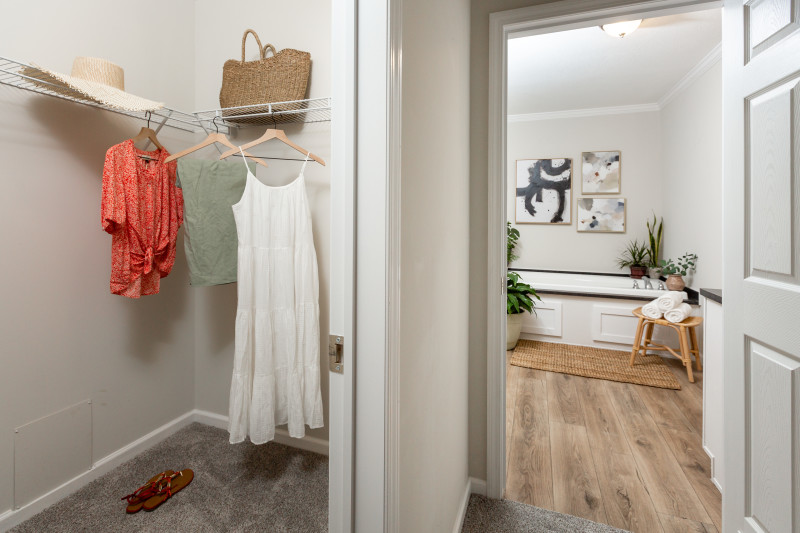 There is a view of part of the split walk-in closet with clothes hung up, a pair of shoes, a purse, and a hat. The bathroom view is off to the right side with a tub in view.