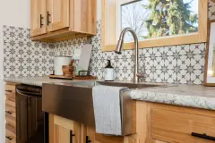 View is of a kitchen window in a manufactured home. There’s a classic starburst pattern backsplash in tan and white. The cabinets are a warm yellowish wood and black bar pulls and there’s a farm sink in the center that is stainless steel.