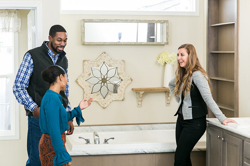 Couple and female home consultant stand in the primary bathroom of a manufactured home with window above the bathtub, shelves built in next to tub and vanity in beige colors.
