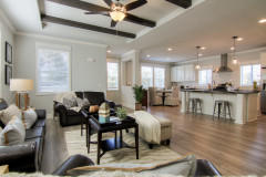 The home has an open floor plan with the living room furnished with a black leather sectional and a wooden chair with a gray throw over it and a large island with metal chairs and white cabinets in the kitchen in the background.