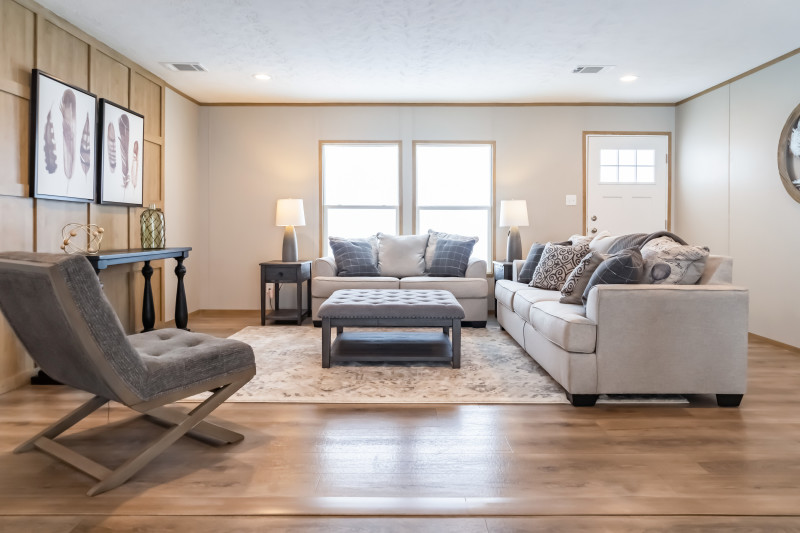 View of manufactured home living room with light coming through. Room is white and light brown with neutral colored furniture.