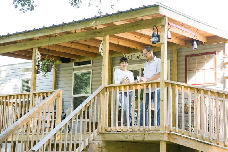 Woman and man stand on the wooden porch of their manufactured home with light gray siding, with wooden stairs.