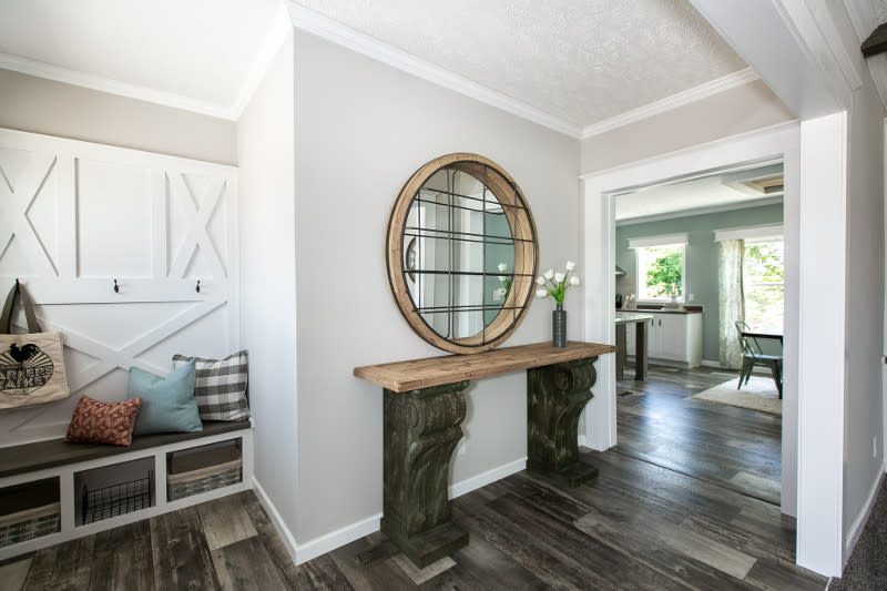 Entryway of a manufactured home with white wood built-in drop zone with coat cooks, bench and cubbies, beige walls and dark wood floor with a wooden table and round mirror.