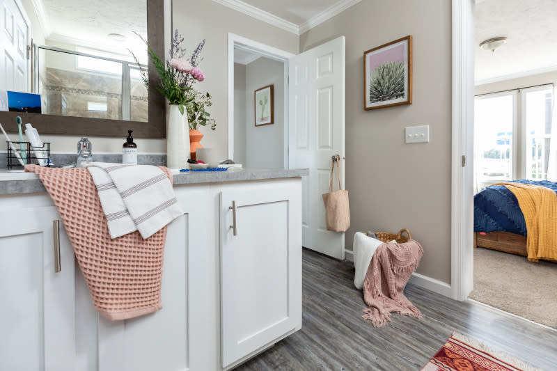 Primary bathroom of a manufactured home with white vanity with gray counters with toiletries, flowers and towels, with a door leading to the hallway and a door to the bedroom.