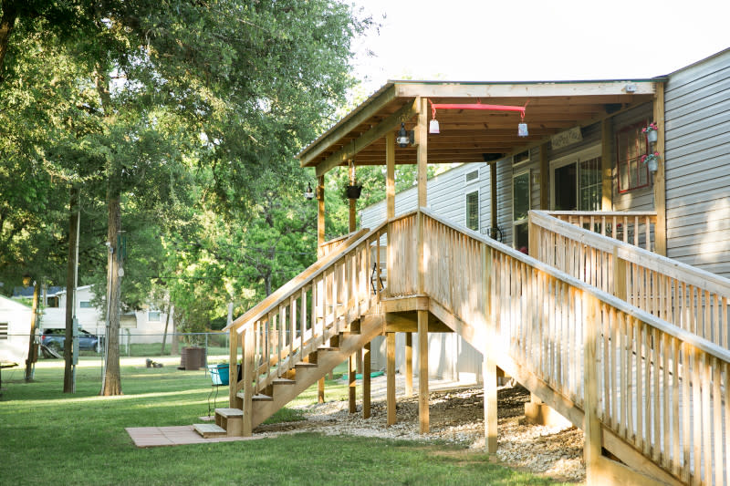 Light wood, covered front porch on a gray manufactured home with stairs in front and a ramp to the right, with trees in the background.