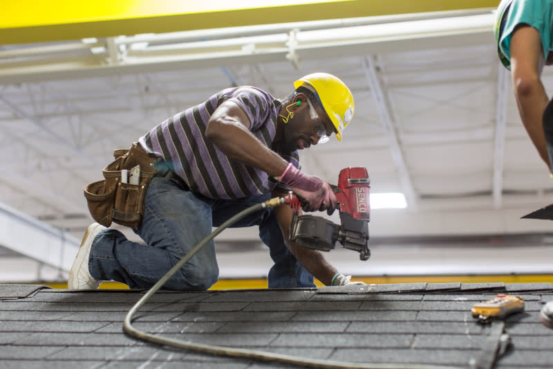 Man wearing T-shirt, jeans, sneakers, hard hat and safety glasses kneels on the roof of a manufactured home with a nail gun.