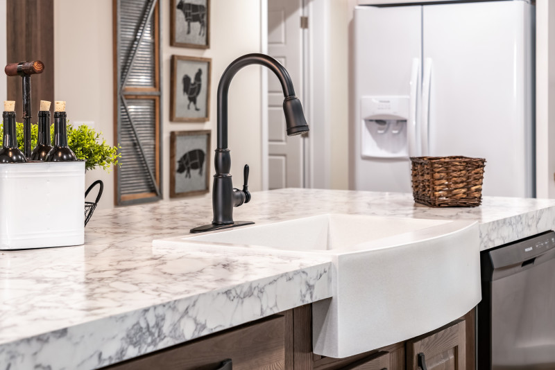 There’s a white sink in a kitchen island with a dark brushed brass faucet head. The countertop is a white-marbled style.