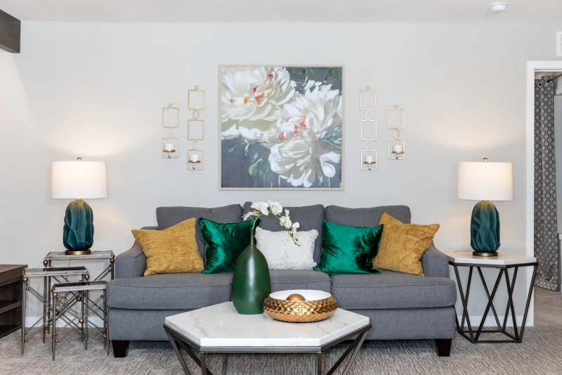 A close-up of a gray couch with yellow and emerald green throw pillows, metal and quartz style coffee and end table and a floral print hanging on the wall.