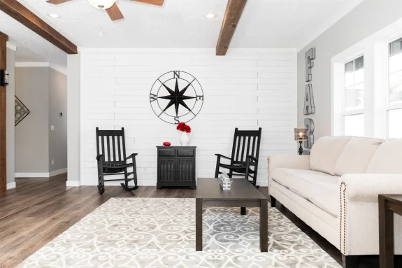 The [model name] features a living room with ceiling beams and a shiplap accent wall.