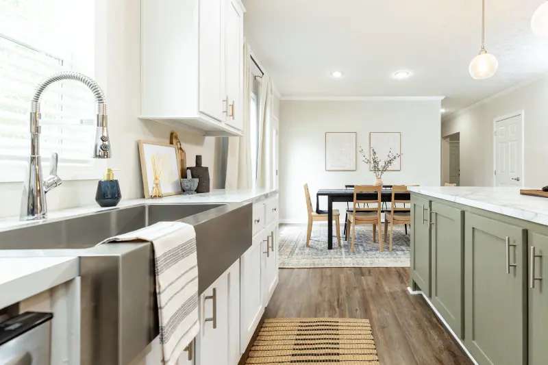 Manufactured home kitchen with white cabinets, sage green island and weathered wood floor, with white walls and a dining room in the background.