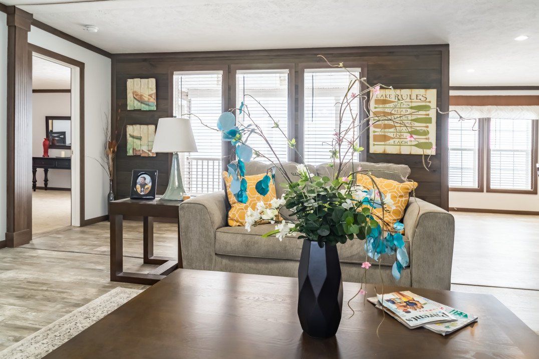 View of the living room in the Turner model manufactured home with a coffee table, gray couch, and three large windows casting light into the room from behind. 