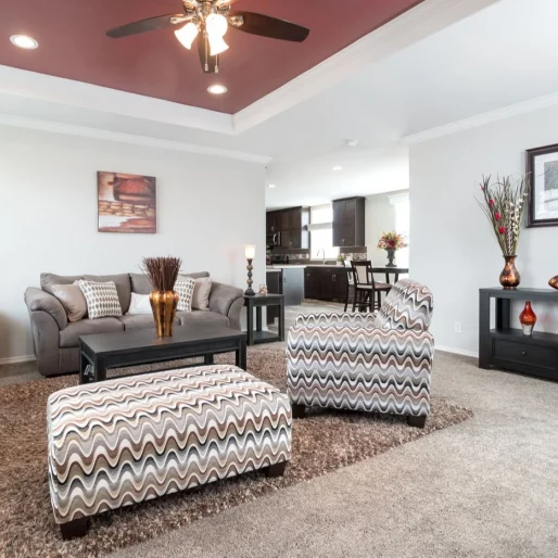 The spacious living room of [model name] has a recessed ceiling with a fan and molding, and leads into the kitchen.