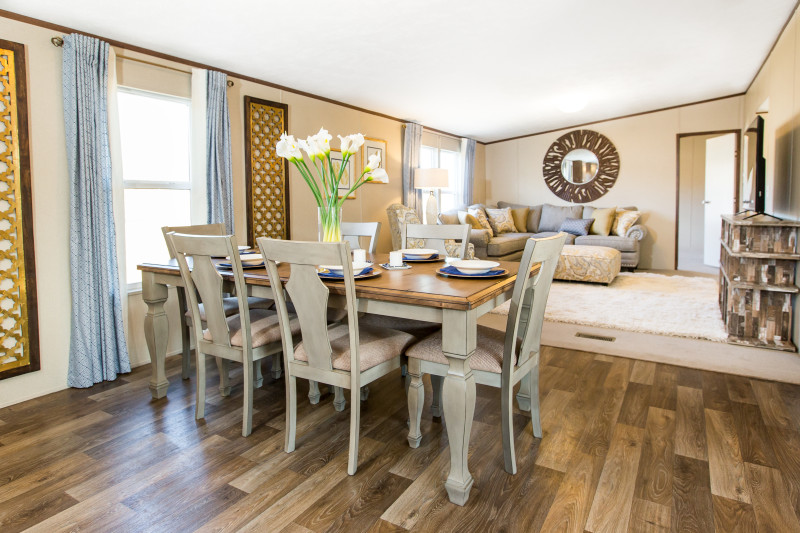 There’s a table with sage green chairs and flowers in a vase on it. The dining area is covered in hard wood flooring. The living room is in the back of the view with some neutral furniture.