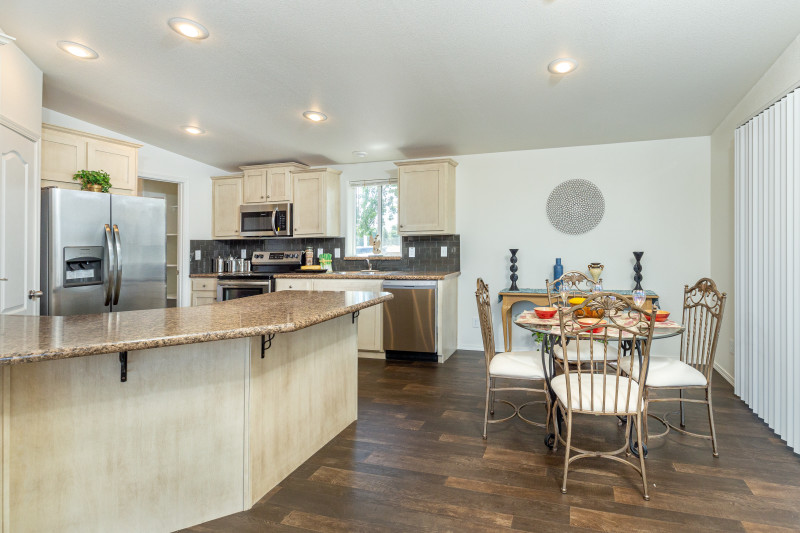 There’s a kitchen that has a dining area to the right with a table set in it. To the left there’s a kitchen with breakfast bar, light colored cabinetry, and stainless-steel appliances. Room is a yellowish white with hardwood floors.