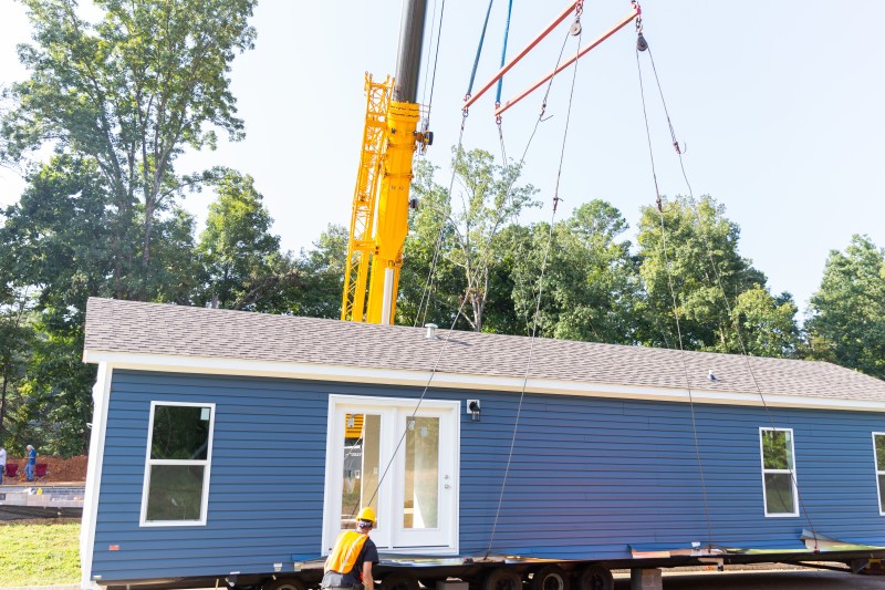 Blue manufactured home with gray roof and white trim being delivered to a home site with a crane in the background and a man in a hard hat in the front.