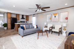 Living room of the Balance manufactured home with neutral modern couch, chairs, coffee table, rug and ceiling fan, with the kitchen in the background.