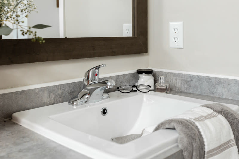 Sink in bathroom of a manufactured home with a gray countertop, white and gray towels hanging from it, glasses and containers next to it and a dark-wood framed vanity mirror.