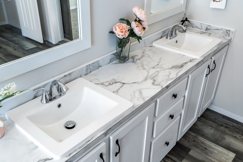 Bathroom of a manufactured home with marble style countertops, 2 white sinks with silver faucets, white cabinets, walls and mirrors, multicolored wood floor and pink flowers on the counter.
