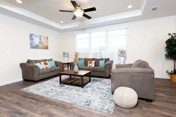 The [model name] features a living room with a tray ceiling and large triple windows.