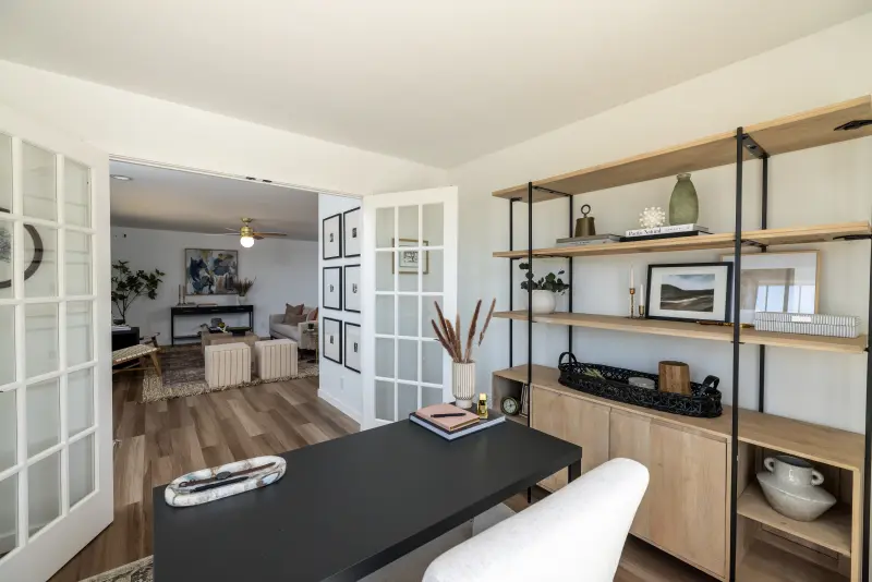 French glass paned doors are open to an office with a black desk and wooden open shelves.