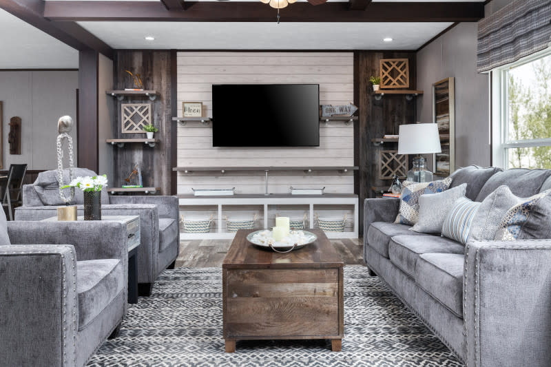 Direct view of the living room in a manufactured home. Accent wall has a tv and décor on either side of it. It has crème colored shiplap and wood panels. The furniture is gray couches and a brown coffee table. There are also wooden beam across the ceiling.