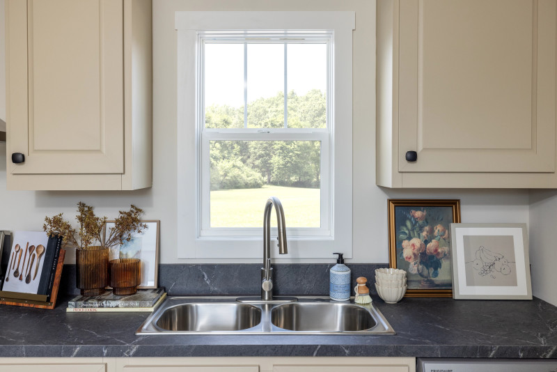 Clayton manufactured home kitchen with cream cabinets, dark countertops, stainless steel sink and a window behind it, with decor on the counters